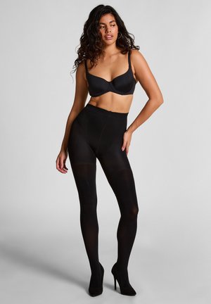 Collants opaques noirs avec une taille haute, associés à un soutien-gorge noir. Le mannequin pose en talons hauts, mettant en valeur la coupe et la texture du vêtement.