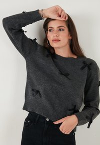 Grijze gebreide trui met zwarte strikaccenten op de mouwen. De stof is zacht, met een relaxte pasvorm en een ribbelrand. Combineren met donkere jeans.