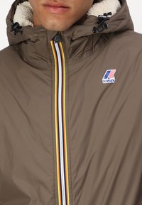 Brown waterproof jacket with a white fuzzy lining, featuring a multi-colored striped zipper and K-Way logo on the chest.