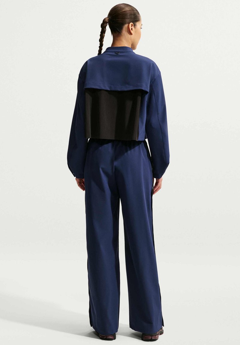 Frau mit geflochtenem Haar, die eine marineblaue Jacke und Hose trägt, Rückansicht mit einem plissierten schwarzen Einsatz an der Jacke.