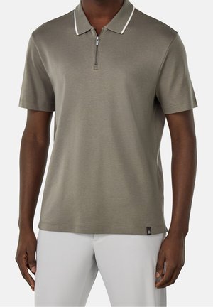 Homme portant un polo taupe manches courtes avec col et fermeture éclair bordés de blanc, assorti à un pantalon gris clair, debout devant un fond blanc.