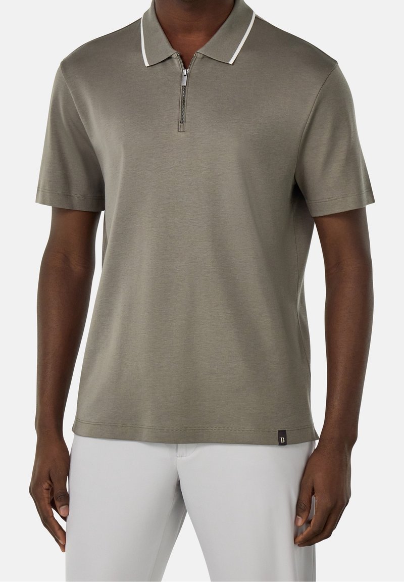 Homme portant un polo taupe manches courtes avec col et fermeture éclair bordés de blanc, assorti à un pantalon gris clair, debout devant un fond blanc.