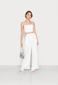 Top cropped branco com alças finas e tecido texturizado, combinado com calças largas brancas. Os acessórios incluem uma mala com padrão de zebra e sandálias brancas.