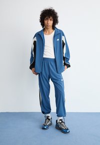 Blue sports tracksuit made of smooth fabric, featuring a zippered jacket with black and white accents and matching pants with elastic waistband.