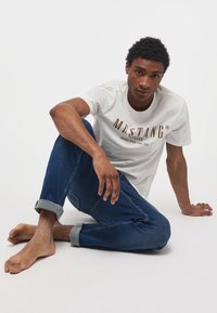 Tricou alb cu logo-ul "MUSTANG" în culoare maro, asortat cu blugi din denim albastri cu mansete rulate. Modelul stă desculț, într-o poziție casual, pe un fundal simplu.