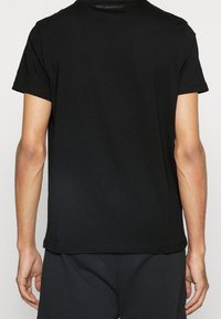 Muž oblečený v jednoduše černém tričku s krátkým rukávem a černých kalhotách, viděn zadu, s textem "Karl Lagerfeld" poblíž límce.