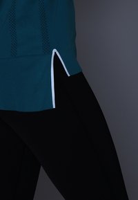 Türkises Sportoberteil mit kurzen Seiten und weißen Akzenten, kombiniert mit schwarzen Leggings. Der Stoff hat eine strukturierte Oberfläche und verfügt über Netzdetails.