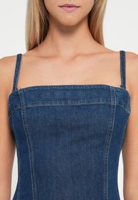 Denim jurk met dunne bandjes, vierkante halslijn en aansluitende corsage. Heeft zichtbare stiksels en een effen donkerblauwe kleur. Soepele textuur.