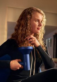 Asciugacapelli Philips con finitura blu opaco, beccuccio a forma di imbuto, impugnatura ergonomica, modello mostrato in uso con capelli mossi, illuminazione soffusa.