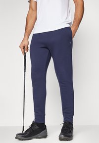 Tumed sinised dressipüksid pehmest kangast, kitsas lõige, elastse vöökohaga, küljetaskute ja väikese logo detailiga reiel.