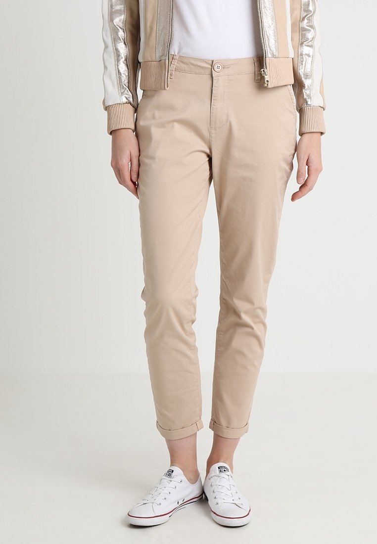 Chinos beige slim fit roulés aux chevilles, portés avec des baskets blanches basses, un top blanc et une veste bomber beige clair et argentée.