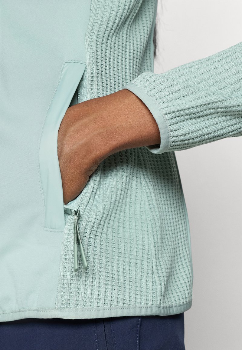 Helleblaue Jacke mit einem strukturierten Strickmuster, seitlicher Reißverschlusstasche und gerippten Bündchen. Elegantes Design mit glatten Stoffakzenten.