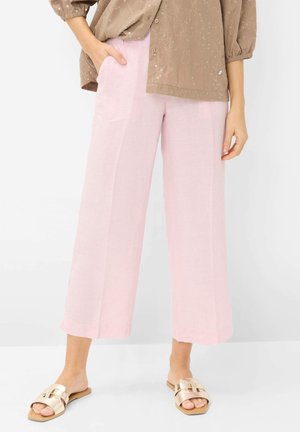 Vrouw draagt lichtroze wijd uitlopende, cropped broek, beige geborduurde blouse en gouden platte slippers, staat met één hand in de zak.