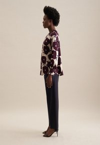 Blusa a stampa floreale in crema e bordeaux, abbinata a pantaloni blu scuro. La parte superiore presenta una vestibilità ampia e maniche lunghe con dettagli a bottoni.