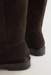 Cizme de gleznă din suede maro, cu vârf rotunjit, fermoar pe lateral și talpă din cauciuc cu nervuri. Detaliile cusute de-a lungul cusăturilor adaugă textură.