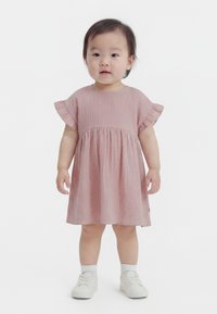 Niña pequeña de pie, vestida con un vestido de manga corta color rosa claro con mangas con volantes, calcetines blancos y zapatillas blancas sobre un fondo blanco.