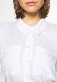 Camicia a righe bianche con colletto, due tasche sul petto e bottoni lucidi. Tessuto morbido e leggero.