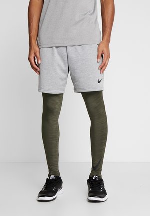 Graue Sportshorts kombiniert mit olivgrünen Leggings. Die Shorts verfügen über ein schwarzes Nike-Logo. Schwarze Sportschuhe vervollständigen das Outfit.