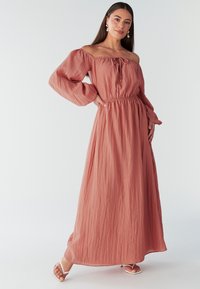 Robe maxi en rouille doux, décolletée avec des manches bouffantes, taille cintrée, texture fluide et ourlet longueur cheville. Associée à des sandales à lanières.