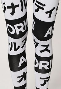 Černobílé legíny s výrazným textem a vzory, vyrobené z elastického materiálu. Design zahrnuje více logotypů v různých orientacích.