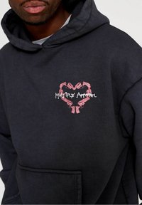 Felpa nera con cappuccio caratterizzata da un design a cuore rosa realizzato con pistole, con il testo "Multiply Apparel" in bianco sottostante. Texture morbida, tasca frontale.