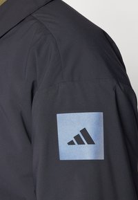 Juodas švarkas su lygiu paviršiumi, matomomis siūlėmis ir mėlynu kvadrato logotipu su trimis juodomis juostelėmis ant rankovės.