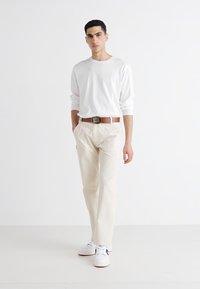 Hvit skjorte med lange ermer, beige bukser, brun belte med metallspenne, og hvite sneakers. Modell står med hendene i lommene.