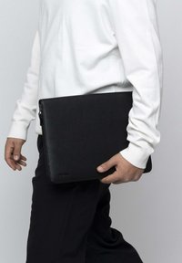 Black leather laptop case with a textured finish, rectangular shape, and top zipper. Held by a person wearing a white long-sleeve shirt.