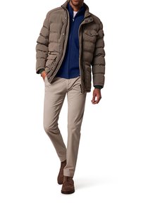 Doudoune marron avec fermeture éclair et poches avant. Sous-vêtement en polaire marine superposé, associé à un pantalon beige et des chaussures en daim marron.