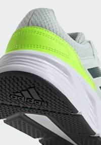 Chaussure de sport avec un dessus gris et un accent vert vif. La semelle est blanche avec un profil de traction noir texturé et le logo Adidas.