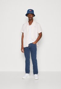 Kortermet, lys rosa skjorte med knapper over en hvit tank topp, parret med blå jeans og en blå bøttehatt med logo.