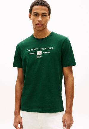 Giovane uomo che indossa una T-shirt verde scuro Tommy Hilfiger con scritte e logo bianchi, abbinata a pantaloni chiari, su uno sfondo semplice.