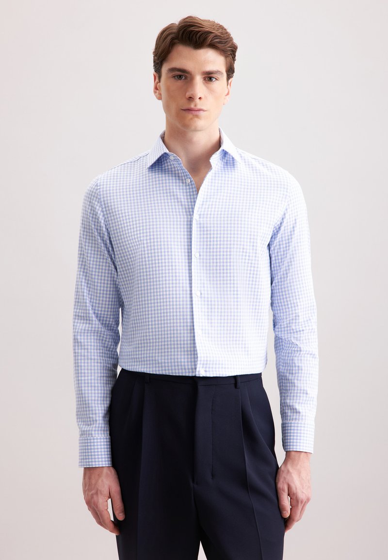 Camicia a scacchi blu chiaro e bianca, maniche lunghe, colletto botton-down, indossata con pantaloni navy. Tessuto morbido, design aderente.