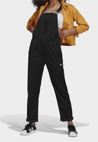 Černé montérky vyrobené z pevné látky, s nastavovacími popruhy, přední kapsou, bočními patentkami a zkrácenými, ohrnutými nohavicemi. V kombinaci s bílým tílkem.