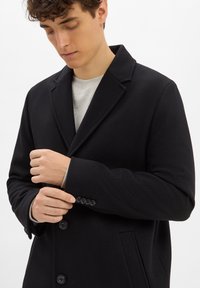 Cappotto lungo in lana nera con colletto a scialle, bottoni frontali e una texture liscia. Presenta maniche aderenti con dettagli di bottoni.