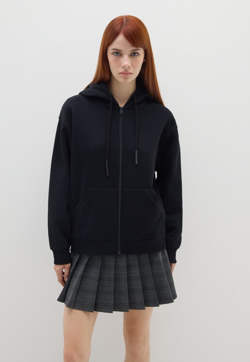 Sudadera negra con cremallera, bolsillo frontal y cordones ajustables, combinada con una falda de cuadros en negro y gris con pliegues.