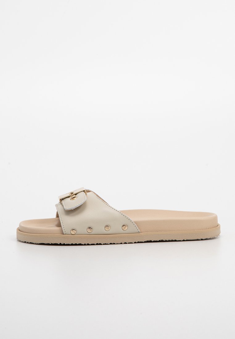 Beige Riemchensandale mit breitem Riemen, goldener Schnallendetails und dekorativen Nieten an der Seite. Glatte Ledertextur, flache Sohle.