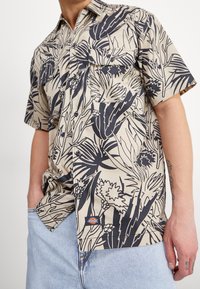 Koszula zapinana na guziki z krótkim rękawem w jasnym beżu, w nawiasie granatowe wzory roślinne, z dwiema kieszeniami na piersi i małą naszywką z logo w prawym dolnym rogu.