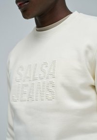 Krema barve pulover s kapuco z embossed logotipom "SALSA JEANS", okrogel izrez in mehak material. Ima rebraste manšete in spodnji rob.