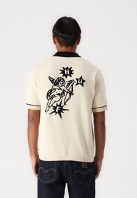 Strick-Polohemd in Creme mit schwarzen Akzenten. Auf der Rückseite befindet sich ein großes schwarzes Grafikmuster eines Cherubins und Sterne. Kurze Ärmel und lässige Passform.
