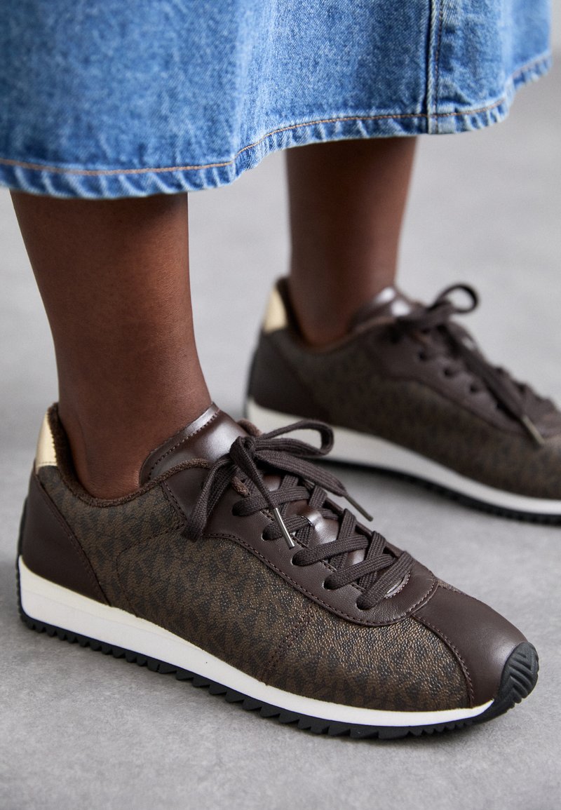 Baskets marron avec une tige en textile à motif, des accents en cuir et une semelle blanche. Les lacets et le détail de l'étiquette ajoutent du contraste.