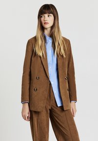 Blazer doppiopetto in velluto a coste marrone con due bottoni, abbinato a una camicia azzurra chiara con bottoni, maniche lunghe e vestibilità rilassata.