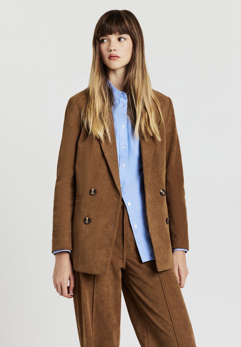 Blazer doppiopetto in velluto a coste marrone con due bottoni, abbinato a una camicia azzurra chiara con bottoni, maniche lunghe e vestibilità rilassata.