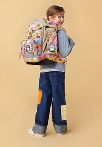 Zaino multicolore a forma arrotondata, con immagini di animali, tasche frontali e base beige. Il bambino indossa jeans con toppe colorate.
