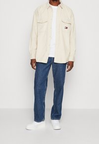 Beige knäppskjorta med två bröstfickor, ihop med blå raka jeans och vita sneakers på en enfärgad bakgrund.