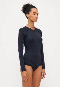 Maglietta da nuoto con zip e maniche lunghe in un blu navy scuro, realizzata in un materiale liscio e elasticizzato. Presenta un design minimalista e una silhouette aderente.