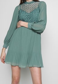 Donna che indossa un mini abito plissé color turchese con lunghe maniche trasparenti e un bodice a motivo reticolare, in piedi di fronte a uno sfondo neutro.