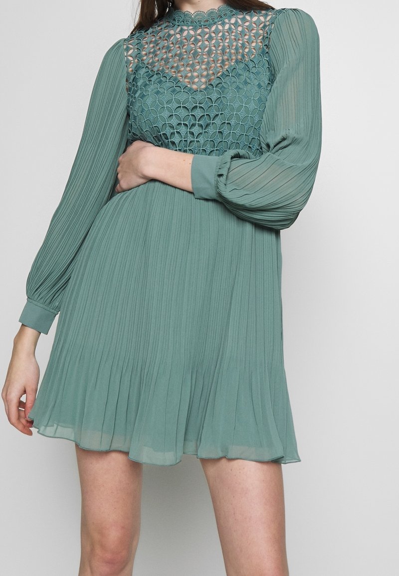 Donna che indossa un mini abito plissé color turchese con lunghe maniche trasparenti e un bodice a motivo reticolare, in piedi di fronte a uno sfondo neutro.