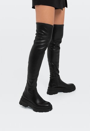 Jambes portant des bottes en cuir noires au-dessus du genou avec des semelles épaisses et des fermetures éclair latérales, assorties à une jupe courte noire sur fond gris.