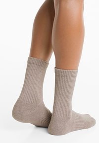 Benen in beige geribbelde sokken tot halverwege het kalf, staand met één voet iets omhoog op een witte achtergrond.